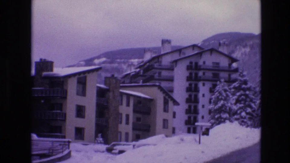 1974 Vail, Colorado 8mm frame showing multi-story apartment buildings with balconies, snow-covered roofs, parked cars, and a wooded mountain backdrop.