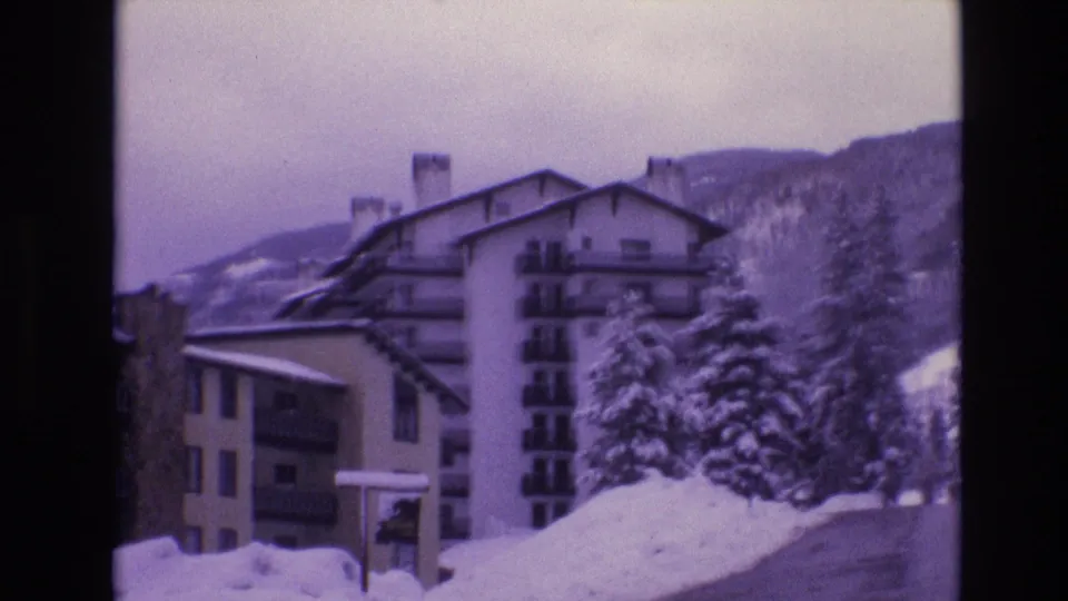 1974 Vail, Colorado 8mm frame showing chalet-style apartment buildings, deep roadside snow, snow-laden evergreen trees, and a forested slope beyond.