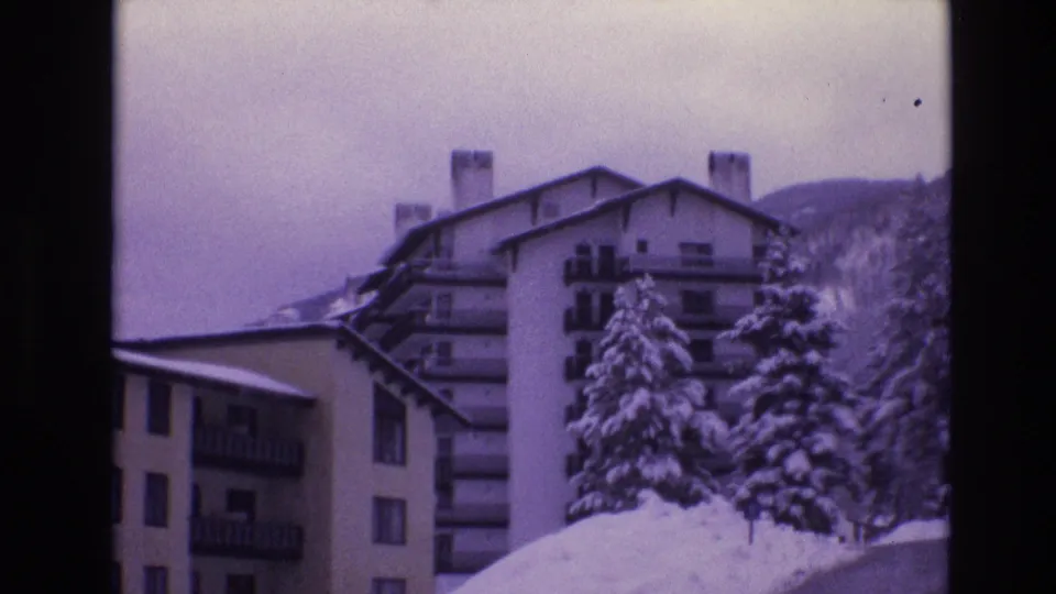 1974 Vail, Colorado 8mm frame showing snow-covered apartment buildings with balconies, a tall evergreen, piled snow, and a wooded mountain background.