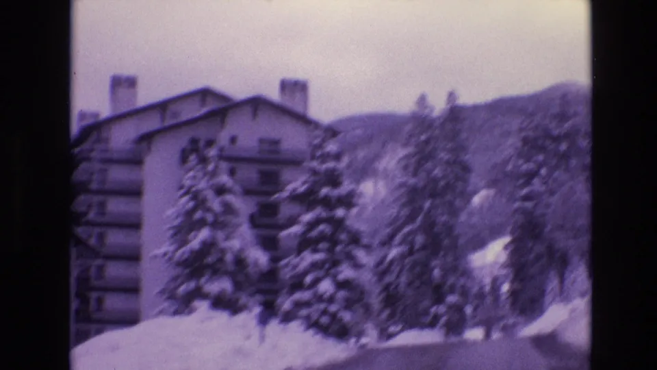 1974 Vail, Colorado home-movie frame showing a multi-story resort building partly obscured by snow-laden pine trees and a mountain slope.