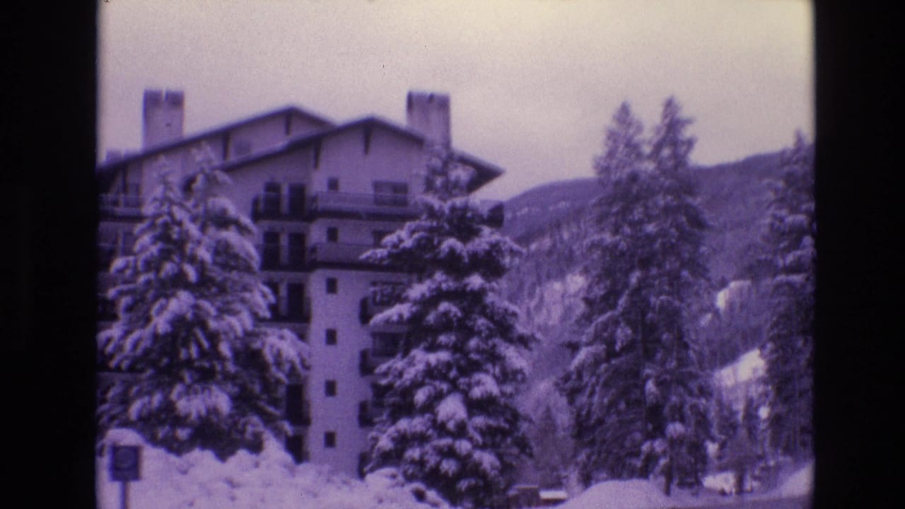 1974 Vail, Colorado frame of a snow-covered hotel exterior with balconies, chimneys, evergreen trees, and mountain terrain behind it.