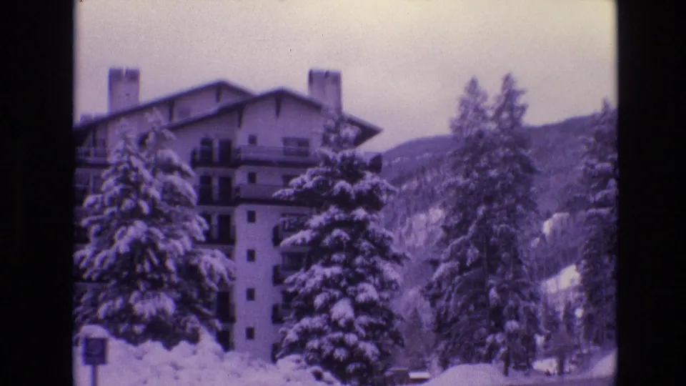 1974 Vail, Colorado frame of a snow-covered hotel exterior with balconies, chimneys, evergreen trees, and mountain terrain behind it.