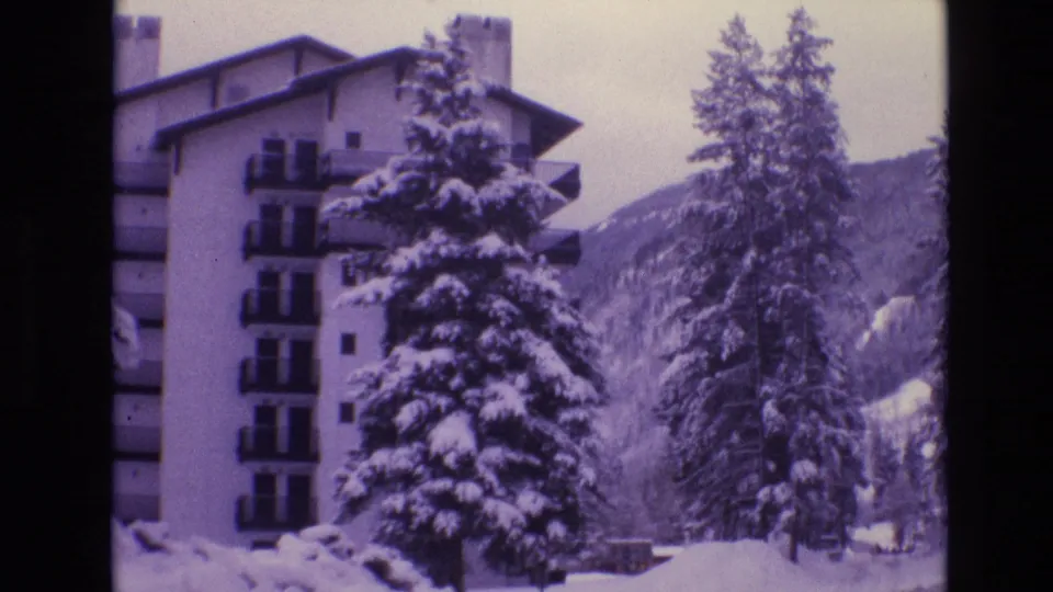 1974 Vail, Colorado home-movie frame showing the side of a multi-story hotel beside tall snow-covered pines and a wooded mountain slope.