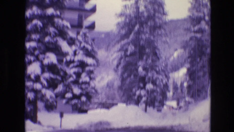 Soft 1974 Vail, Colorado frame of snowy resort grounds with evergreen trees, a partially visible building edge, and a mountain slope in the background.