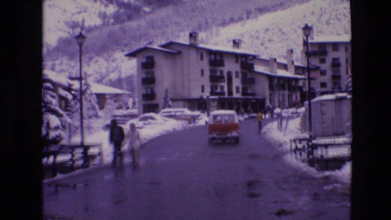 1974 Vail, Colorado street scene with a red van driving away on a wet snow-lined road, bundled pedestrians at left, apartment buildings, lampposts, and snowy mountains beyond.