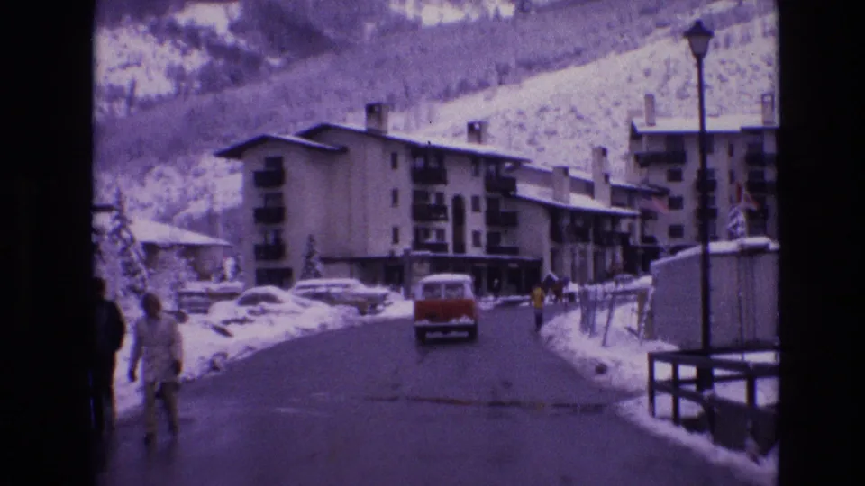 Red vintage van continues down a wet winter road in Vail, Colorado, with pedestrians near the left edge, snow piled along the street, and low-rise buildings against snowy slopes.