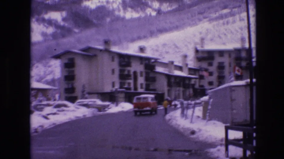 Soft Super 8 frame of a Vail winter street, showing a red van farther down the road, snowbanks, parked cars, apartment buildings with balconies, and a bundled pedestrian near the right edge.