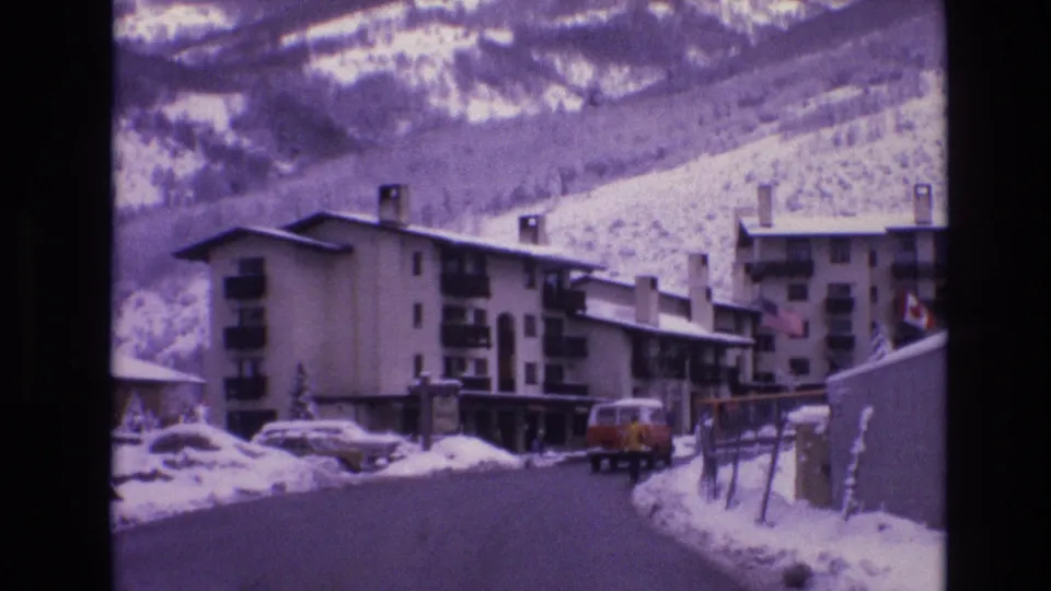 Late frame of the 1974 Vail street view, with the red van receding beside snowbanks, parked cars and apartment buildings lining the road, and snowy mountains rising behind the town.