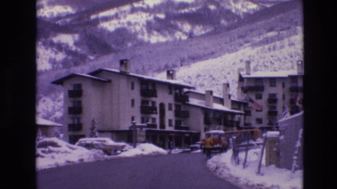 1974 Vail, Colorado home-movie frame showing a snow-lined resort road, multi-story lodges, parked 1970s cars, and snowy mountain slopes in the background.