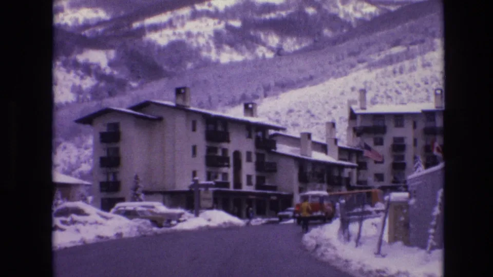 1974 Vail, Colorado home-movie frame showing a snow-lined resort road, multi-story lodges, parked 1970s cars, and snowy mountain slopes in the background.