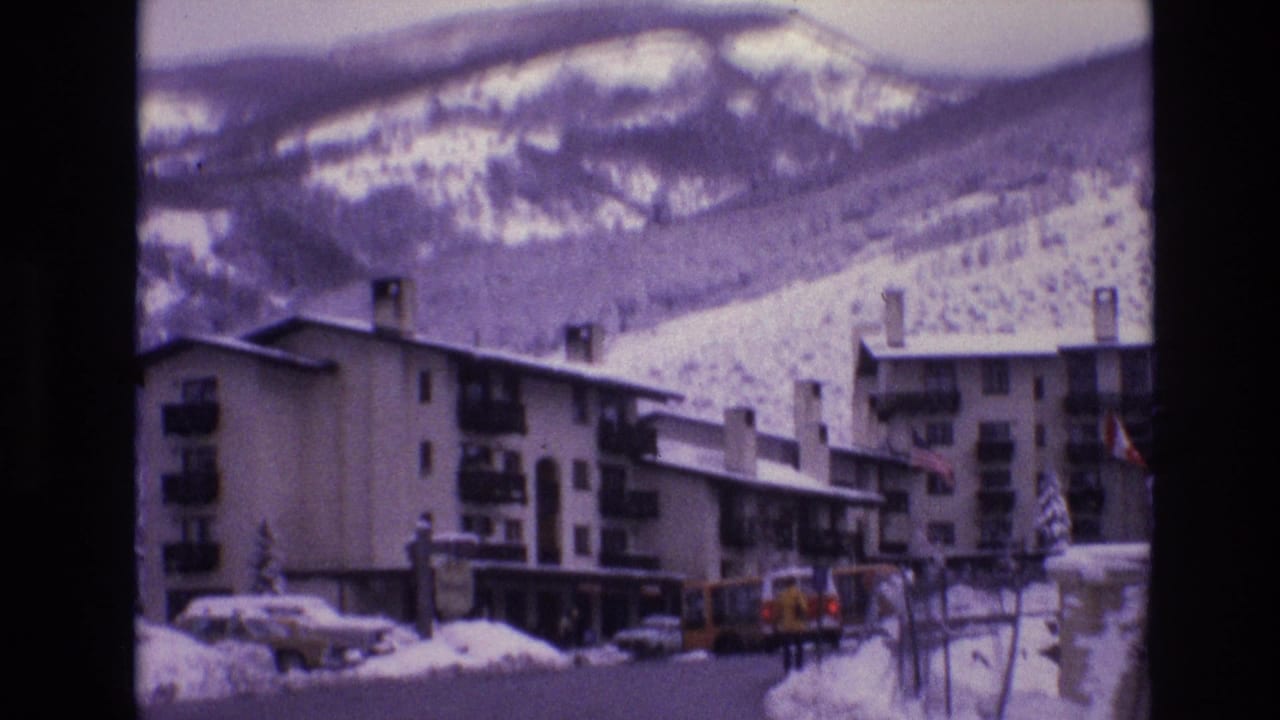 1974 Vail, Colorado frame with ski-lodge buildings, parked cars, heavy roadside snow, and a mountain backdrop seen through a purple-tinted 8mm scan.