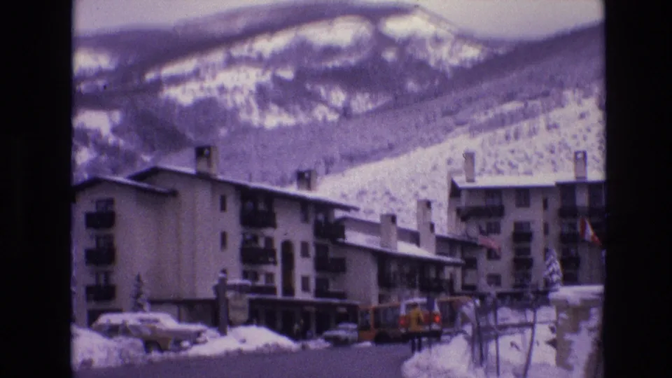 1974 Vail, Colorado frame with ski-lodge buildings, parked cars, heavy roadside snow, and a mountain backdrop seen through a purple-tinted 8mm scan.