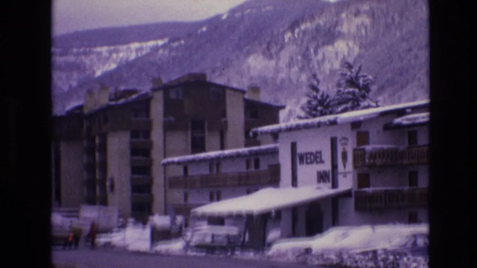 1974 Vail, Colorado home-movie frame showing the Wedel Inn sign on a snow-covered resort building with balconies, parked cars, pedestrians, and mountains behind.