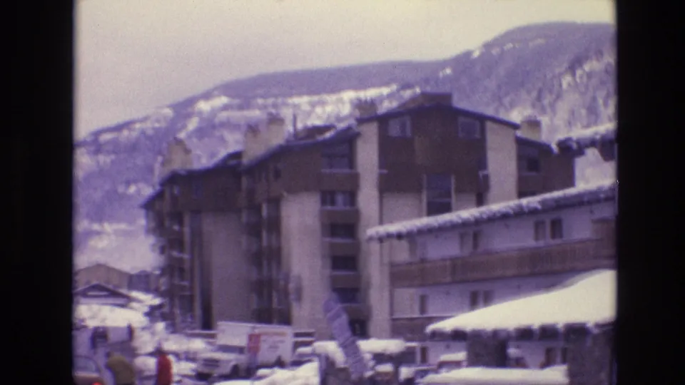 1974 Vail, Colorado frame showing snowy lodge buildings, parked cars, a small person in a red jacket near the street, and mountains beyond the resort area.
