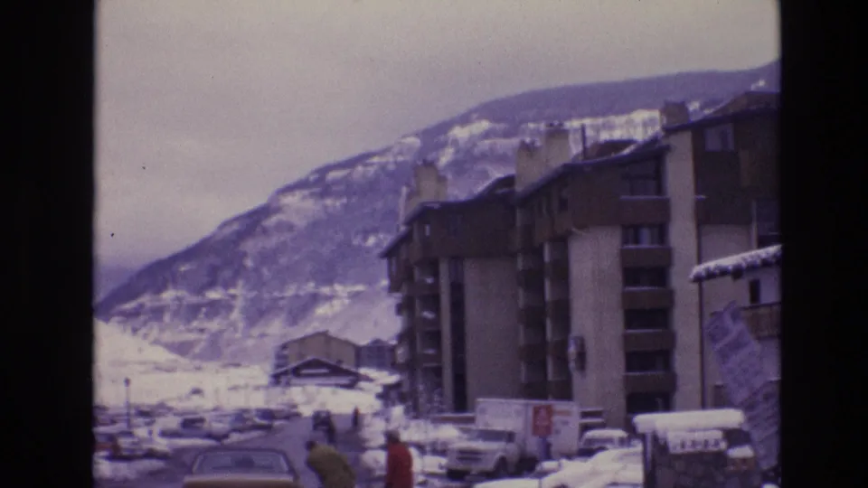 1974 Vail, Colorado frame looking along a snow-covered resort street with lodge buildings, parked cars, a distant pedestrian, and mountains in the background.