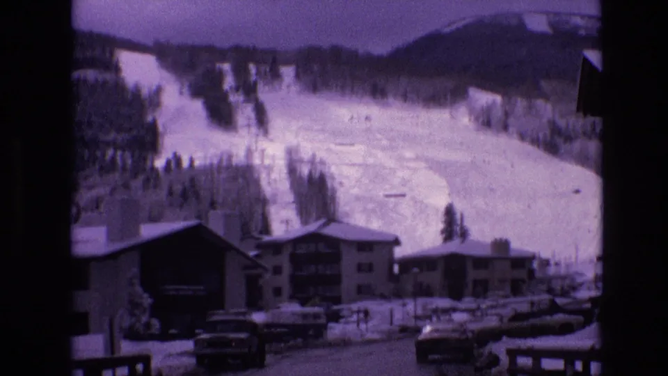 1974 Vail, Colorado frame with a snowy ski slope rising behind lodge buildings, parked cars, fencing, and a winter road in a resort village.