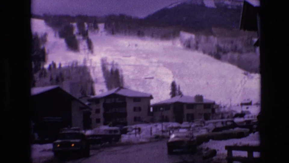 1974 Vail, Colorado home-movie frame showing parked cars and low resort buildings at the base of a broad snowy slope, with visible 8mm grain and soft focus.
