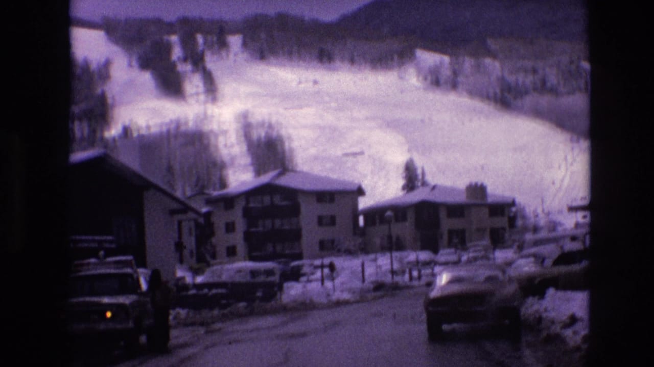 1974 Vail, Colorado home-movie frame showing a snow-covered village street, low-rise ski lodges, parked vintage cars, and white mountain slopes beyond.