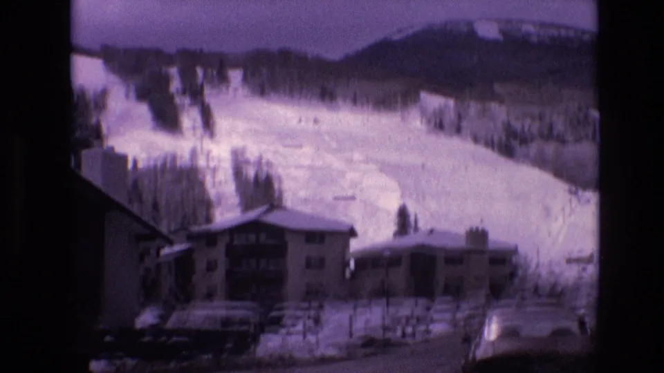 1974 Vail, Colorado frame with a snowy roadside view of ski lodges, parked automobiles, fencing, and broad ski runs on the mountain behind.