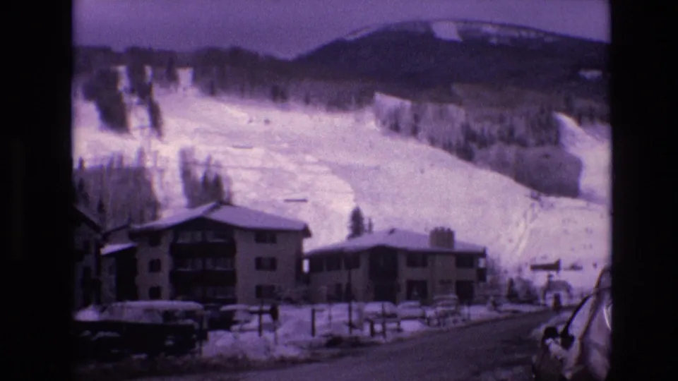 1974 Vail, Colorado home-movie still showing a snow-lined road, lodge structures, parked cars, fencing, and ski terrain rising behind the village.