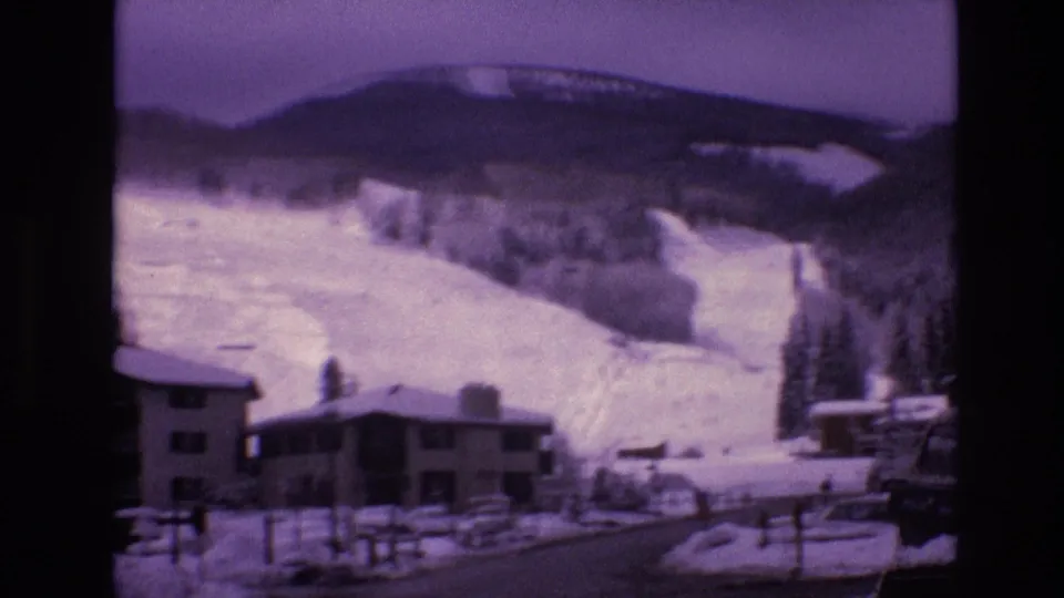 1974 Vail, Colorado frame showing snowy lodge rooftops, a curved village road, fencing, evergreen trees, and ski slopes under a purple-shifted sky.