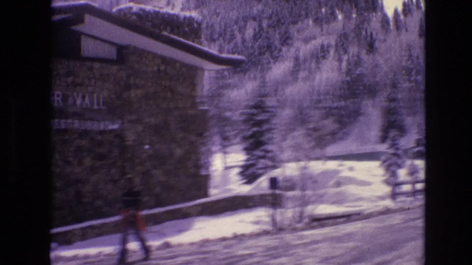 1974 Vail, Colorado home-movie frame with a pedestrian moving past the lodge exterior beside a snow-lined road and evergreens.