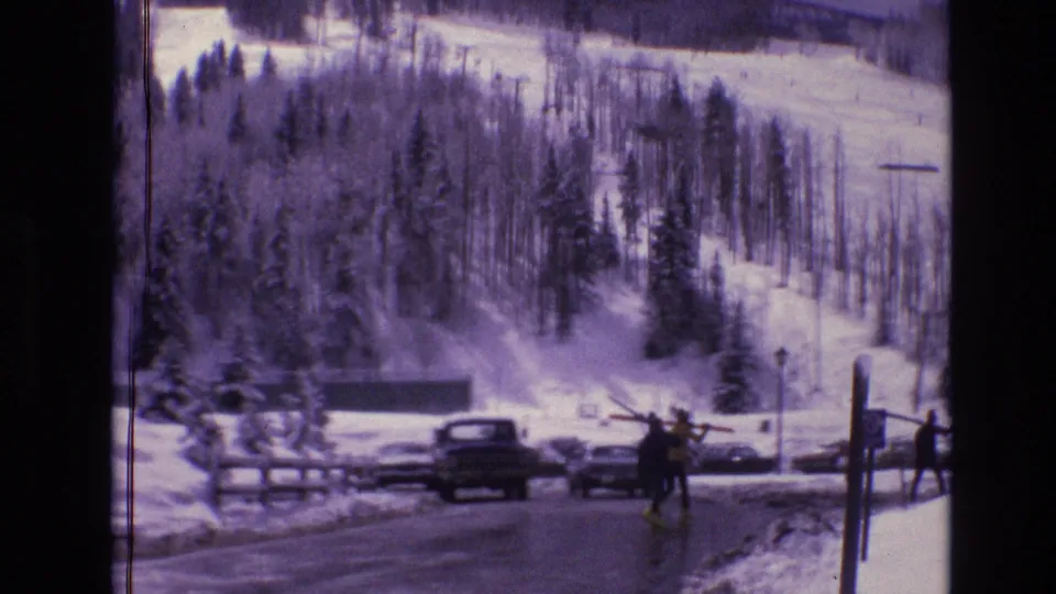 1974 Vail, Colorado frame showing skiers with gear near parked vintage cars at the snowy base area of Vail Mountain.
