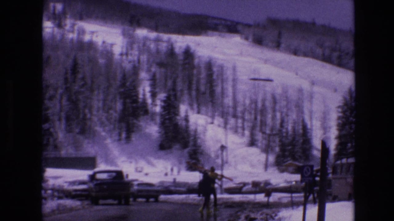 1974 Vail, Colorado 8mm frame showing people with skis in a snowy resort parking area, with parked cars, evergreens, and ski runs behind them.