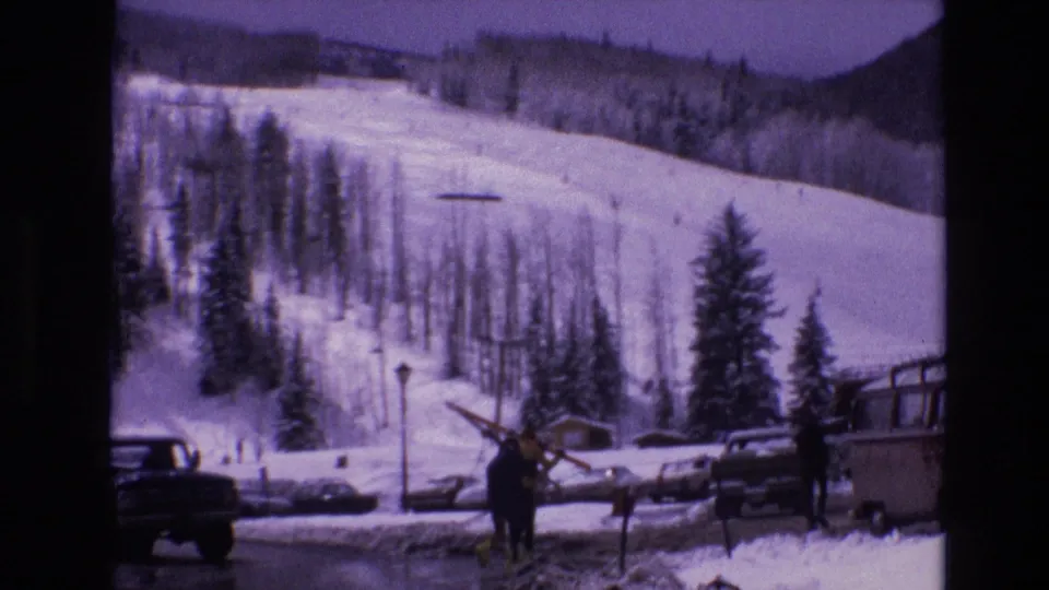 A skier carries equipment through a snow-covered Vail resort parking area, with classic vehicles, a bus or camper, trees, and ski slopes in the background.