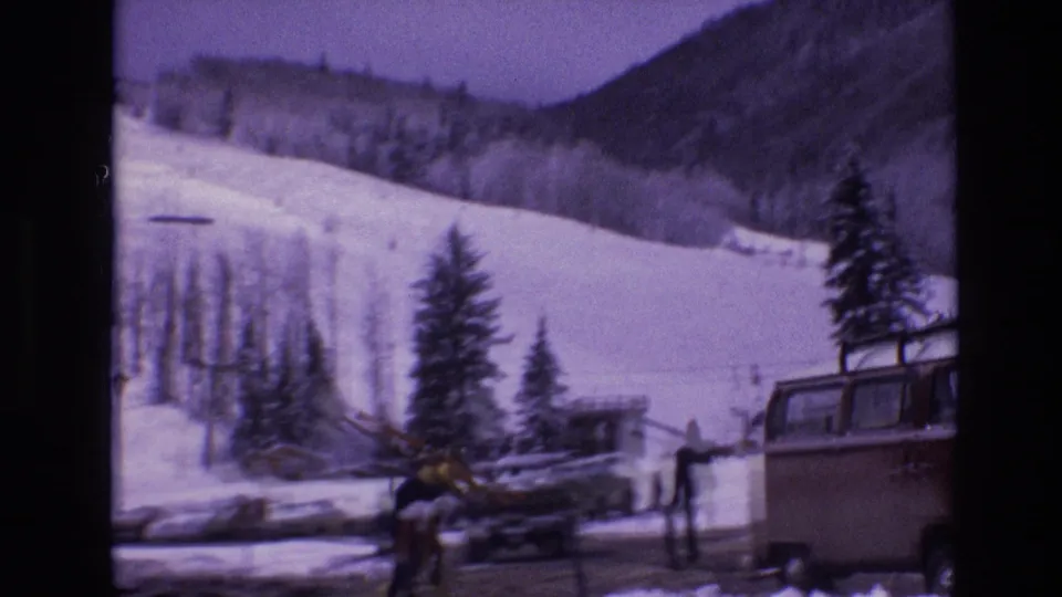 A soft 8mm view of Vail ski slopes and a snowy parking area, with people near the road, parked vehicles, evergreens, and a large bus or camper partly visible.