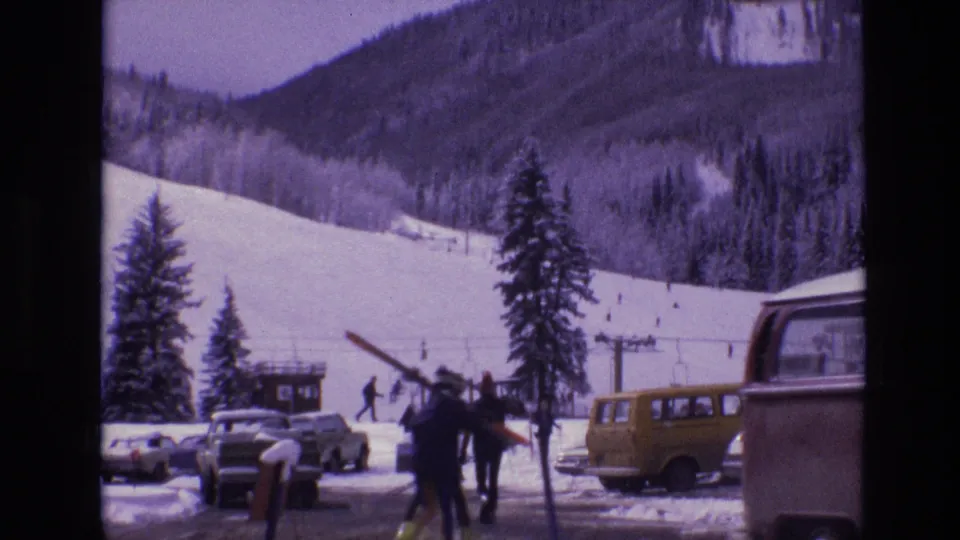 1974 Vail resort scene with skiers carrying gear near a yellow van, parked cars, a large bus or camper, lift structures, and snow-covered mountain slopes.