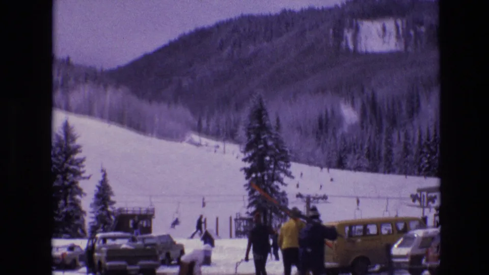 Vail, Colorado ski-resort base area in 1974 with skiers on a snowy slope, people in winter coats, parked cars, and a yellow station wagon in the foreground.