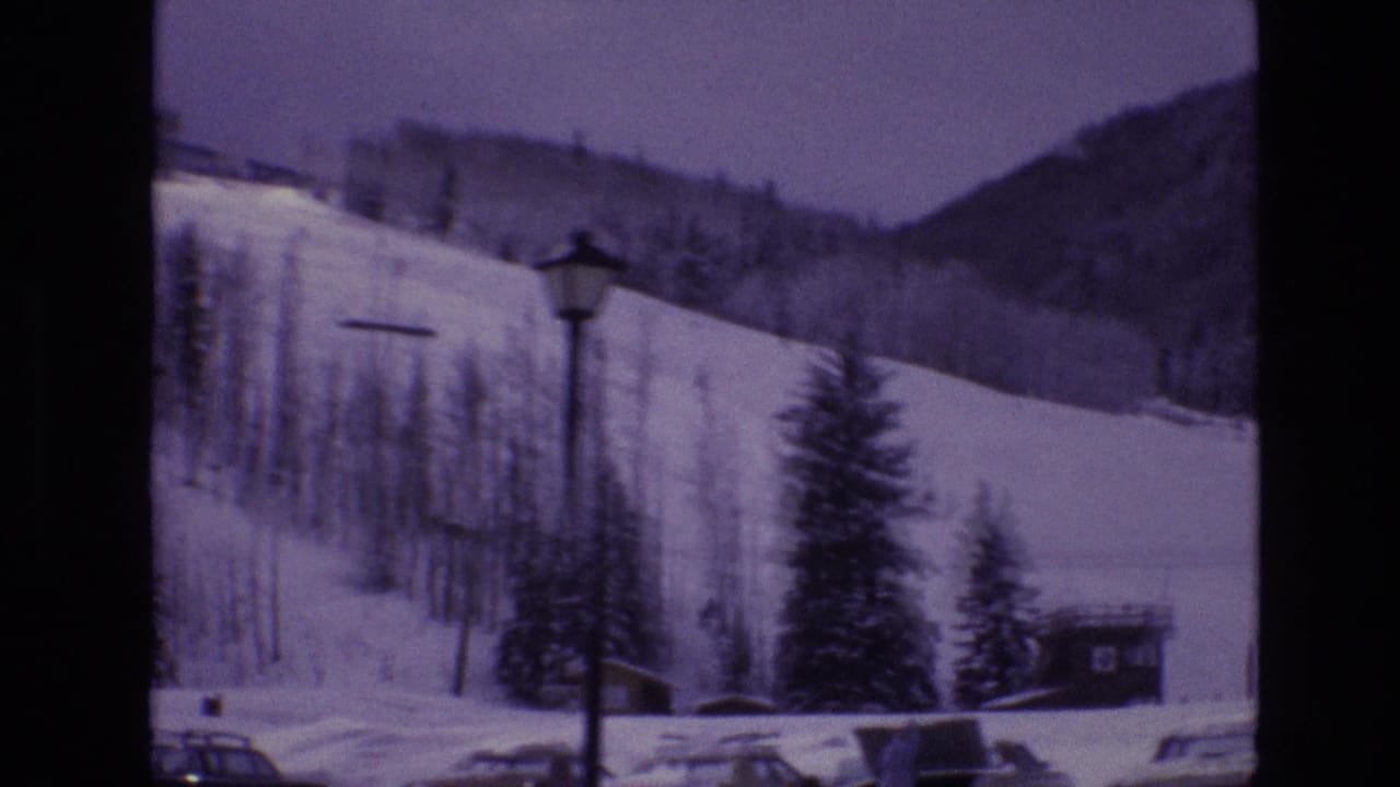 Snowy hillside with ski runs, trees, parked cars, and a lamp post near the resort base.