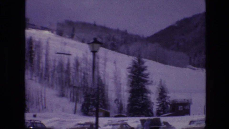 Snow-covered Vail ski terrain in 1974 with a cleared run, leafless trees, evergreens, parked cars, and a lamp post seen from the resort base area.