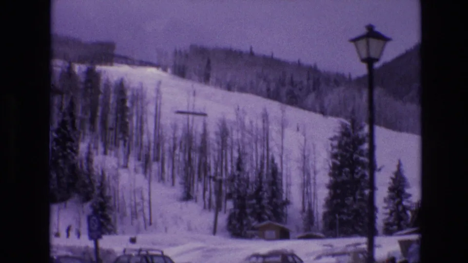 1974 Vail ski-resort view with a snowy run, bare trees, evergreens, parked cars, a small structure, and a tall lamp post near the base.