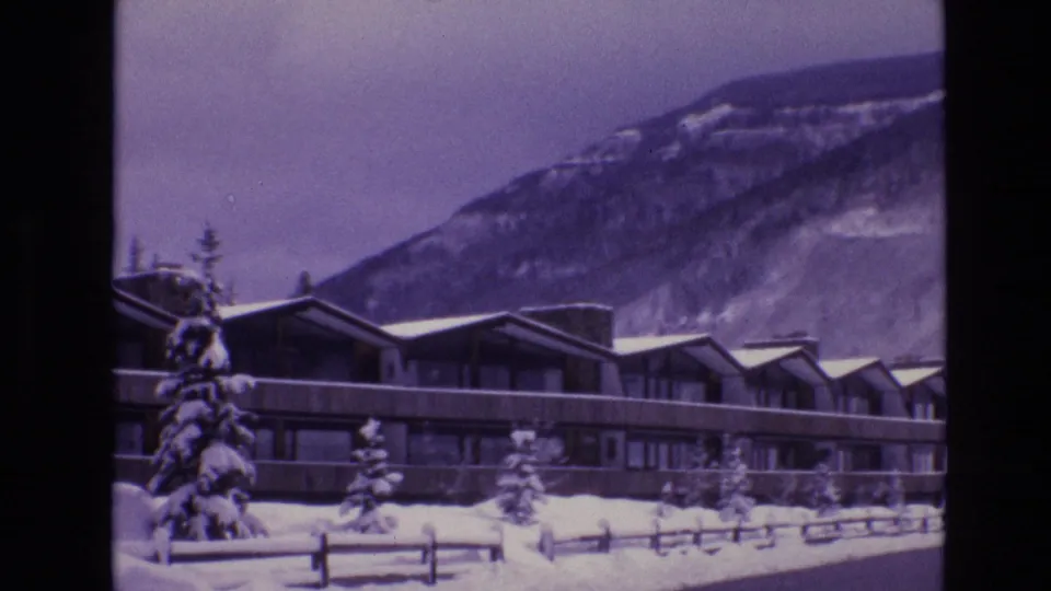 1974 Vail 8mm frame showing a long low lodge exterior, snow-covered roofs, small evergreens, fencing, and a mountain slope beyond.