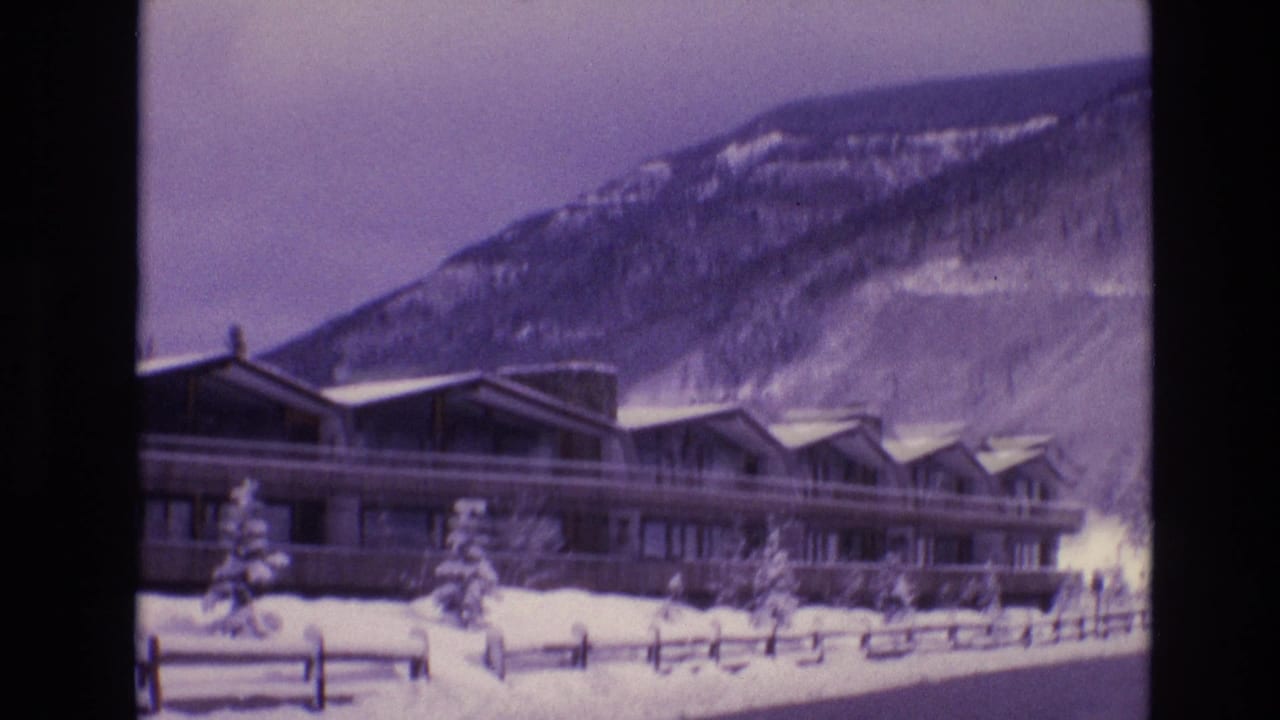 1974 Vail, Colorado 8mm frame showing a long modern lodge with peaked roof sections, snowbanks, evergreens, and a mountain slope behind it.