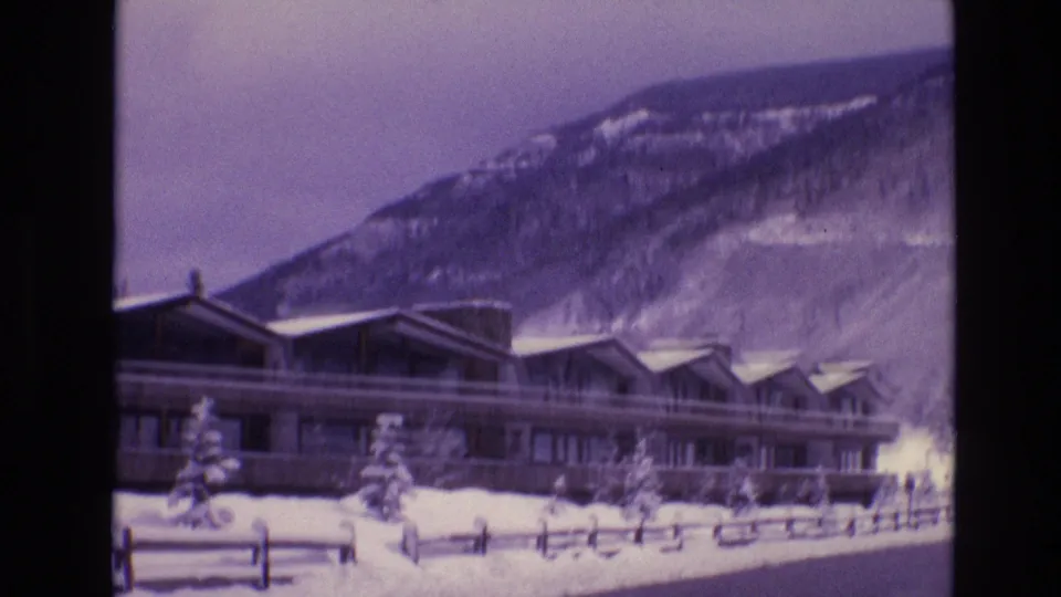 1974 Vail, Colorado 8mm frame showing a long modern lodge with peaked roof sections, snowbanks, evergreens, and a mountain slope behind it.