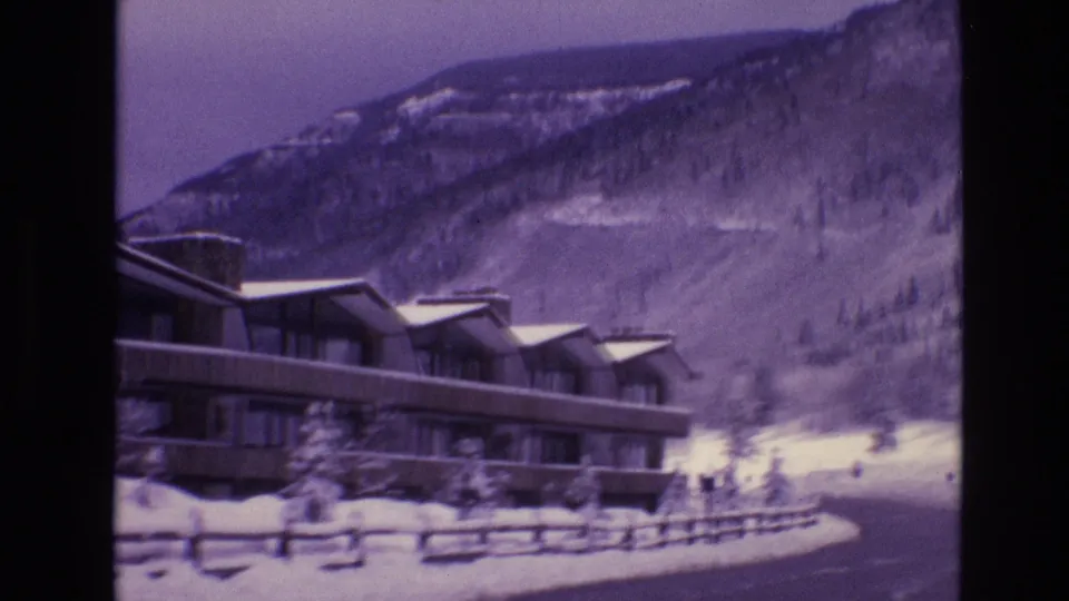 1974 Vail, Colorado 8mm frame showing part of the lodge at left, a split-rail fence, deep snow, a plowed road, and a wooded mountain slope.