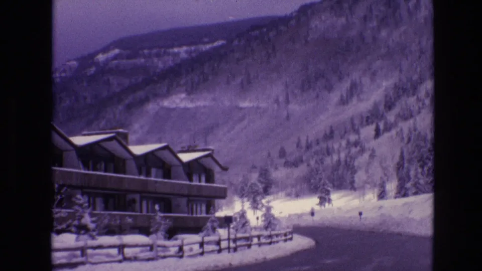 1974 Vail, Colorado 8mm frame with the lodge at lower left, a snow-lined road curving through the foreground, fence rails, evergreens, and a wooded mountain slope.