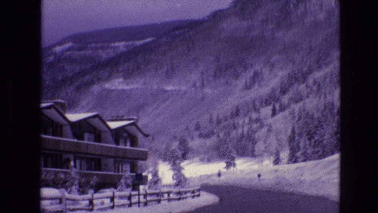 1974 Vail, Colorado home-movie frame showing a snow-covered lodge, roadside fence, winding winter road, evergreen slope, and rugged mountain backdrop.