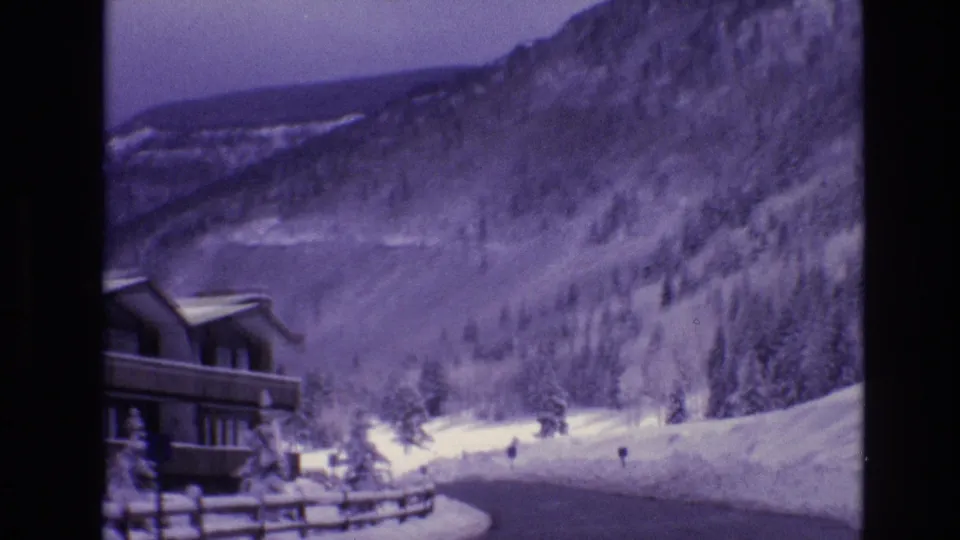 A visitor-shot 1974 Vail resort view frames a lodge at left, a curving snowbound road, distant small figures, evergreen trees, and a steep mountain slope.