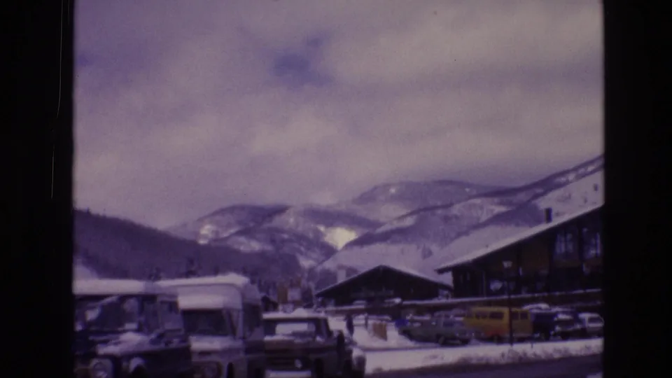 1974 Vail parking-area view with boxy cars and vans in snow, a ski-lodge building, low clouds, and rounded mountain ridges behind the resort.