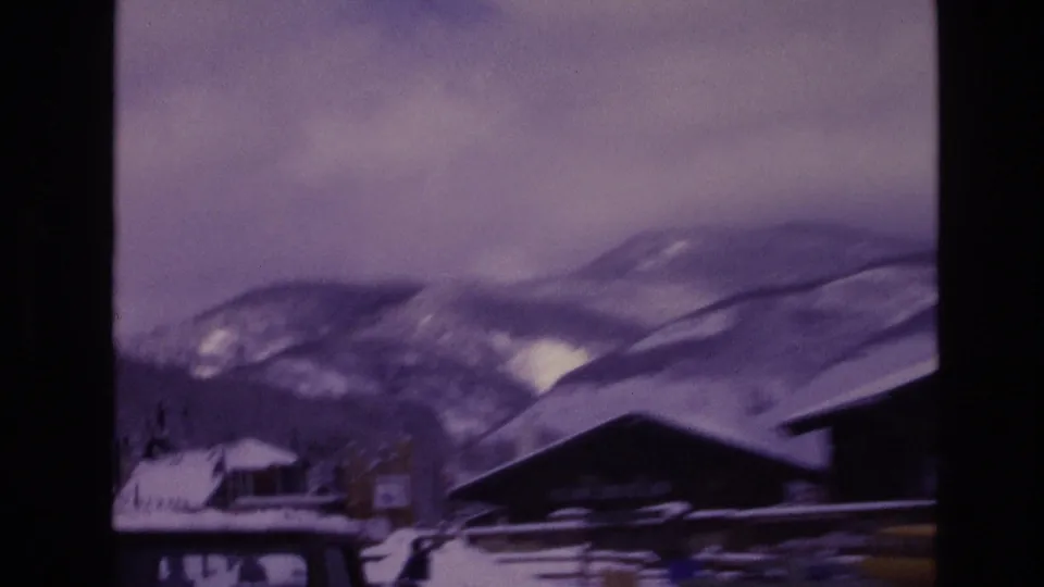 A soft 8mm frame from Vail in 1974 shows a low lodge roofline, snow-covered ground, distant mountain ridges, and a clouded sky with pronounced color shift.