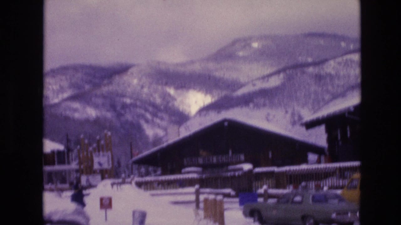 1974 Vail, Colorado 8mm frame showing a dark wooden lodge, snowy ground, parked vehicles, railings, and mountain slopes under an overcast sky.