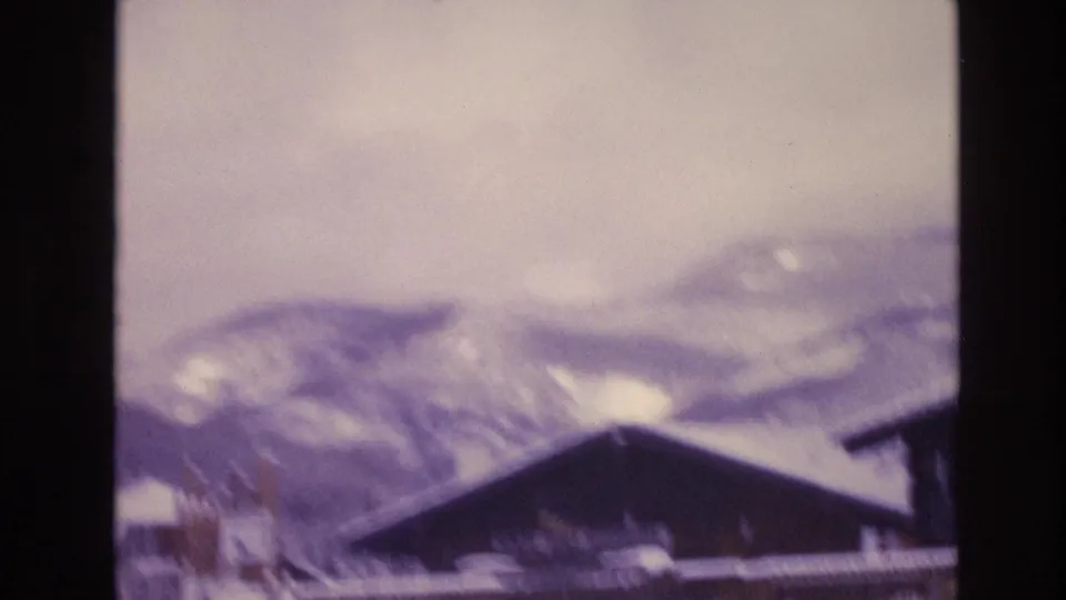 1974 Vail, Colorado 8mm frame with a dark sloped lodge roof, snow-covered mountain backdrop, pale winter sky, and partial neighboring building edge.