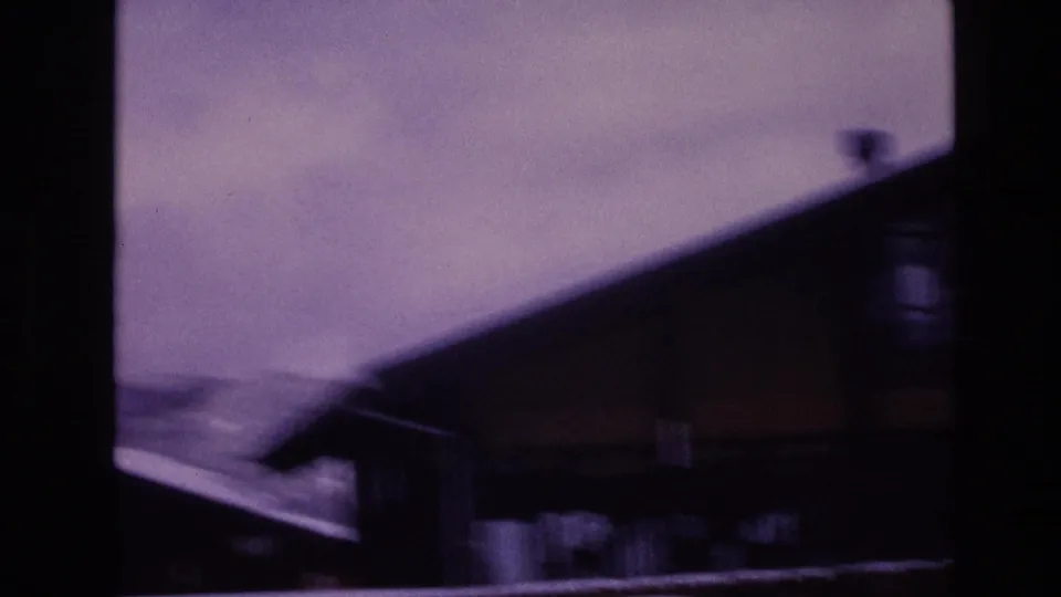 1974 Vail, Colorado 8mm frame with a dark wooden building in the foreground, snow along the roofline, blurred mountain slopes, and pale winter sky.