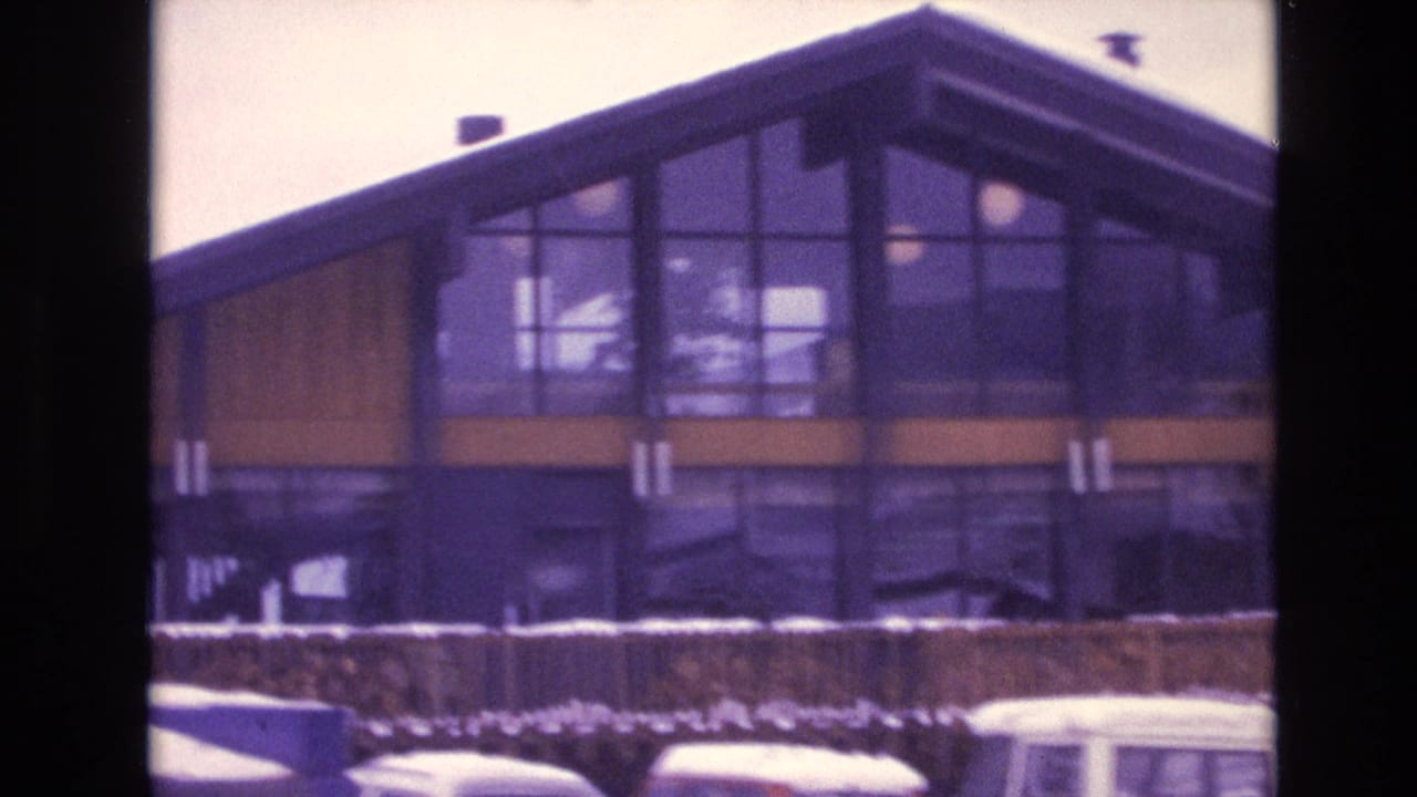 Vail, Colorado 1974 home-movie frame showing a modernist ski lodge with large windows, wood-toned panels, snowbanks, and cars along the foreground.