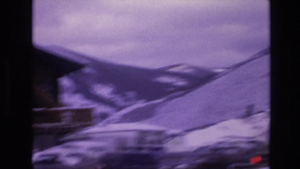 Vail, Colorado 1974 frame with a snowy roadside, blurred lodge roofline, parked vehicles, and dark mountain slopes in the background.