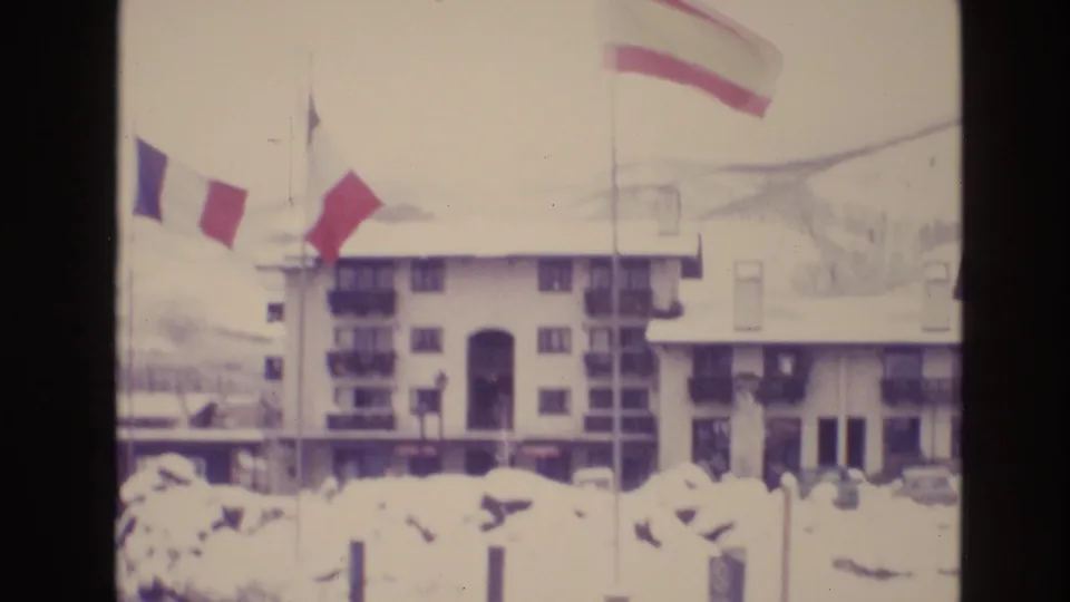 Vail, Colorado 1974 home-movie frame showing a multi-story hotel or lodge building behind snowbanks, with several flags on poles in the foreground.